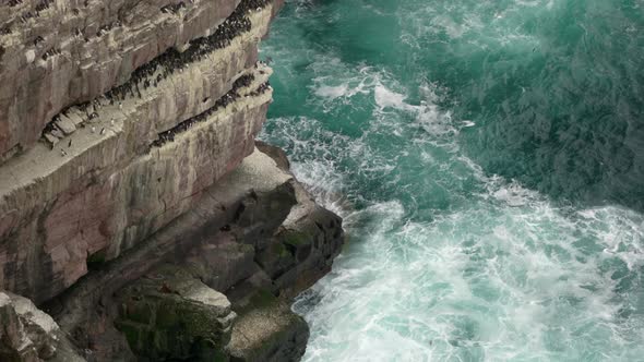 Powerful waves crash over rocks and against a sea cliff in the middle of a beautiful, deep teal ocea