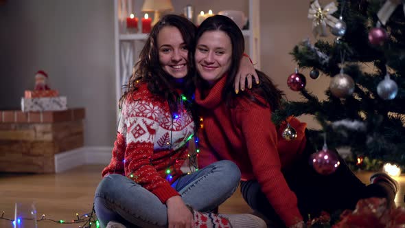 Two Happy Young Caucasian Women Sitting at Christmas Tree Hugging Smiling Looking Away Talking