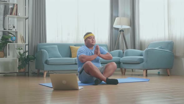 Fat Asian Man On Mat Looking At Laptop And Doing Yoga In Sukhasana Pose At Home