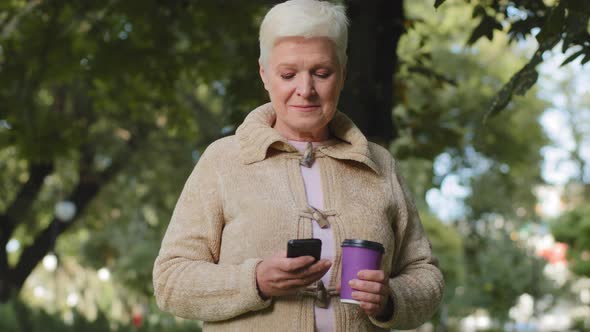 Smiling Attractive Elderly Grayhaired Retired Woman Walking in Park with Plastic Cup of Coffee and