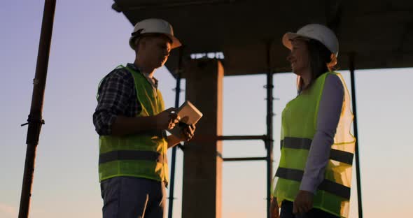 Builders Man with a Tablet and a Woman in White Helmets Shake Hands at Sunset Standing on the Roof