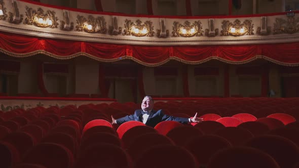 Successful man in suit alone in large auditorium. Actor man is sitting among empty red armchairs