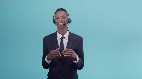 Black Office Man in a Headset Talks Gesticulating with Hands and Staring at the Camera on a Blue