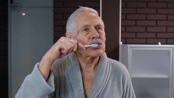 Senior Man Grandfather in Bathrobe Brushing Teeth, Looking Into Mirror. Morning Hygiene at Bathroom