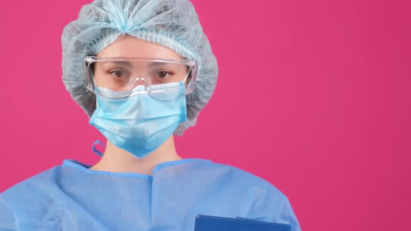 Serious Doctor in a Blue Medical Coat Safety Glasses and Medical Cap Looking at Camera