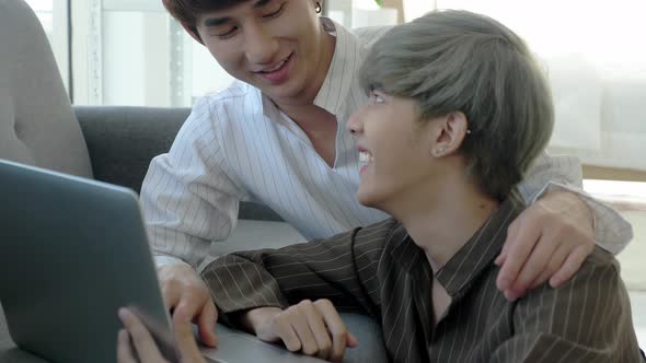 Asian loving gay couple cuddling, using laptop, smiling, making eye contact