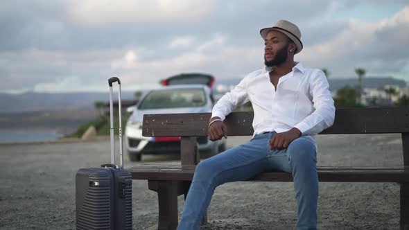 Wide Shot Portrait of Thoughtful African American Gay Man with Trolley Bag Sitting on Bench Looking