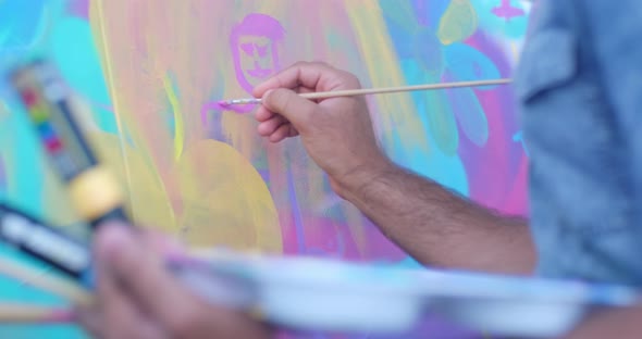 Tight shot of a man painting a stick figure in pink on a big cloth canvas in a public park on a summ