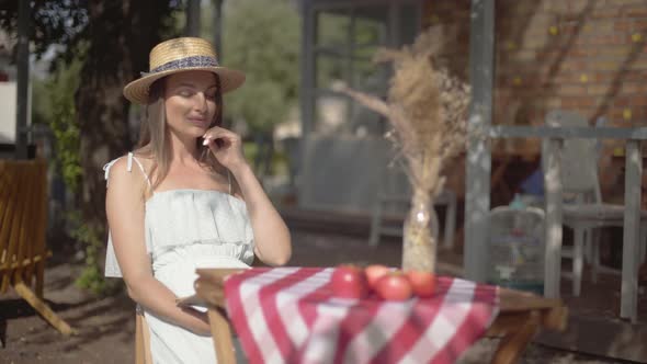 Young Girl in a Straw Hat and White Dress Sitting at the Small Table and Looking at the Camera