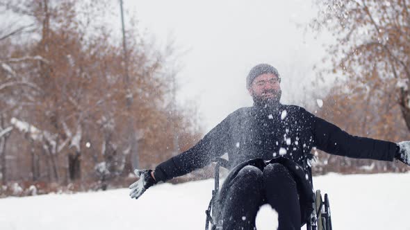 Laughing disabled man in wheelchair throws snow over himself in park, front view. Happy disabled