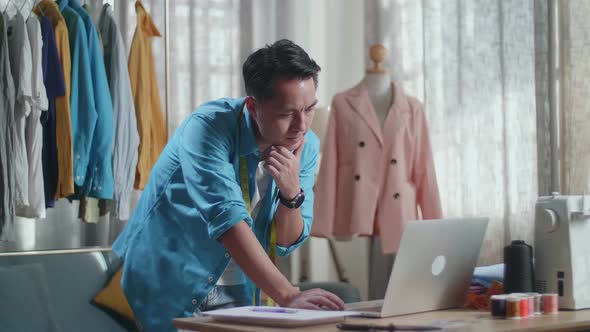 Asian Male Designer Working On A Laptop And Thinking While Designing Clothes In The Studio