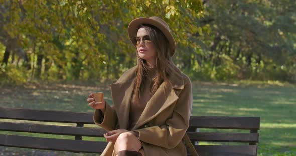 Elegant Caucasian Woman in Brown Hat and Sunglasses Drinking Tea or Coffee As Sitting on the Bench