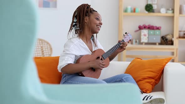 Attractive Young Woman is Playing Guitar While Sitting on Sofa in Apartment Interior Spbi