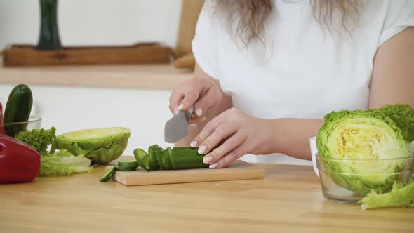 Young Blonde Woman with Curly Hair Cuts a Cucumber Into Rings While Sitting at a Table in the