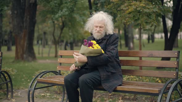 Romantic Senior Caucasian Man Sitting on Bench in Autumn Park Smelling Bouquet of Flowers
