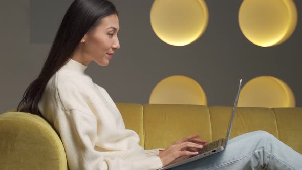 Emotional Young Asian Woman with a Laptop Celebrates Victory on a Yellow Sofa at Home