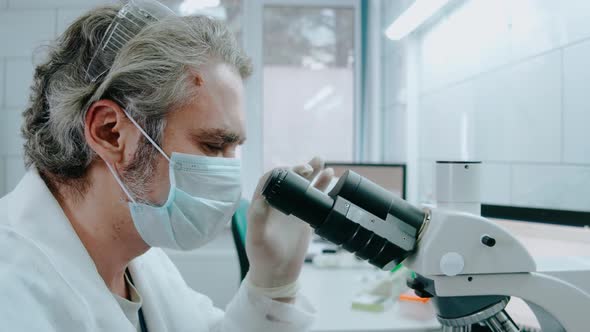 Laboratory Assistant Looks Through a Microscope and Writes Notes in a White Lab