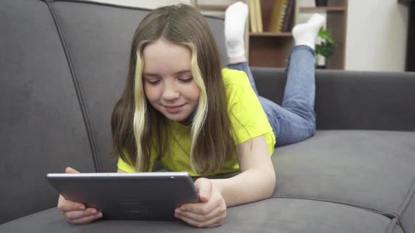 A Little Girl is Smiling While Lying on the Couch with a Tablet