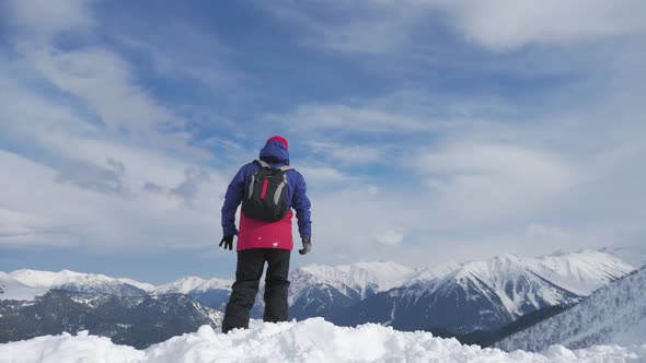 Man with Backpack Standing on Snowy Mountain Top in Winner Pose with Raised Hands Enjoying View