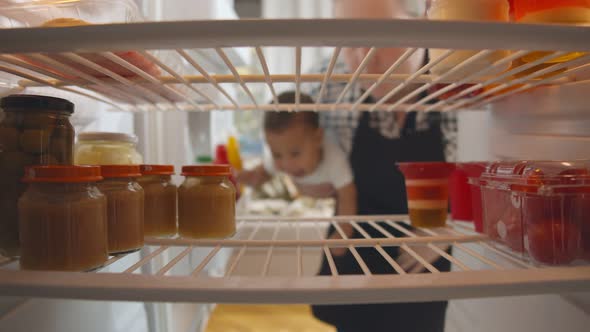 Mother with Baby in Hands Opening Fridge and Taking Glass Jar of Baby Food