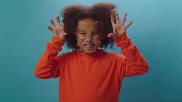 Cute African American girl wearing cosmetic face mask of tiger standing on blue background