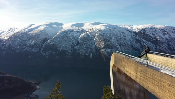 Tourist relaxing on tip of Stegastein viewpoint platform while drone slowlying along side and passin