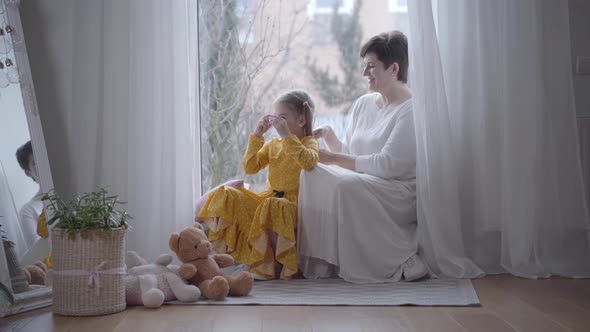 Pretty Positive Girl Playing with Toy As Grandmother Tying Her Hair in Ponytails. Happy Caucasian