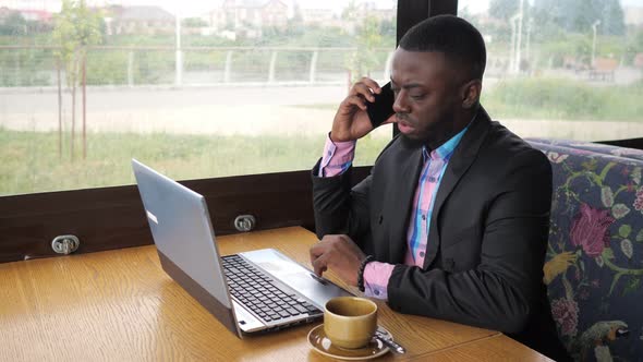 Black Businessman Works on Laptop and Calls Smartphone Sits in Cafe with Coffee