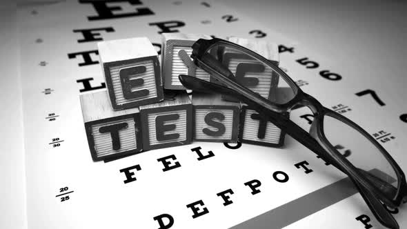 Glasses Falling Next To Blocks Spelling Out Eye Test In Black And White alt