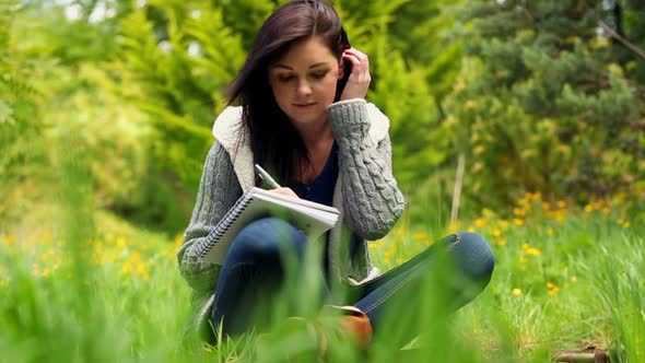 Smiling Young Woman Sitting On Grass Writing On Notepad