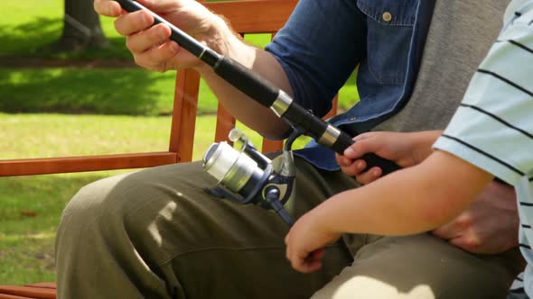 Father Teaching His Young Son How To Fish On A Bench