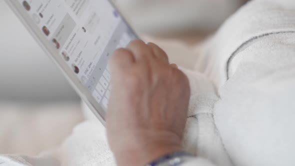 Senior Woman Lying on a Bed in a White Terry Robe Uses a Tablet, Writes a Message in the Messenger