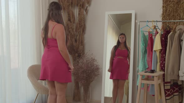 Plump Young Adult Woman in Pink Dress Admiring Appearance Indoors