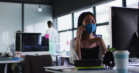 Asian woman wearing face mask talking on smartphone while sitting on her desk at modern office
