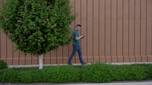 A Young Man Goes with a Phone in His Hand. A Man Walks Near the Building and Carries on