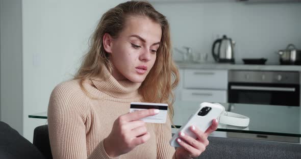 Smiling Young Woman Customer Holding Credit Card and Smartphone Sitting on Couch at Home