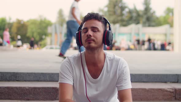 A Young Handsome Man in a White Tshirt and Headphones Sits on the Steps in the City and Listens to