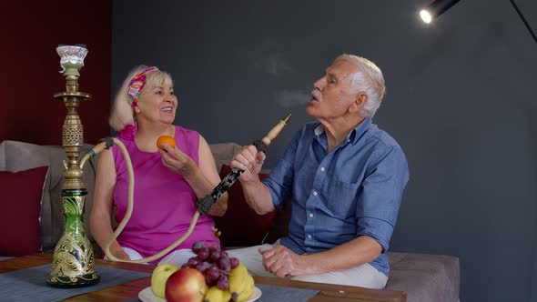 Senior Couple Smoking Hookah Eating Fruits Celebrating Retirement Anniversary in Living Room at Home