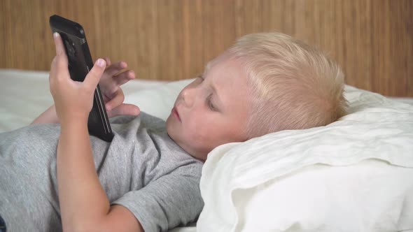 The boy lies on the bed with a smartphone in his hands.