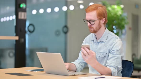 Casual Redhead Man Using Smartphone and Laptop in Office 