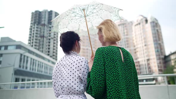 Back View Two Happy Women in Elegant Dresses with Sun Umbrella Standing Outdoors Admiring Urban