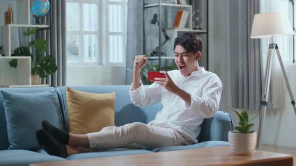 Full Body Of Asian Man Celebrating Winning Game On Smartphone While Lying On Sofa In The Living Room