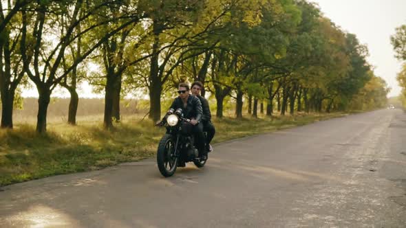 Tracking Shot of Young Cool Couple Riding Motorcycle on Forest Road in Autumn