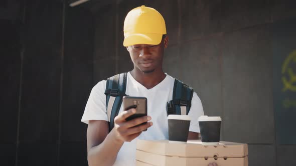 African Delivery Worker Checking His Phone While Carrying a Box with Pizza and