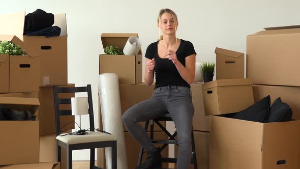 A Happy Moving Woman Sits on a Chair in an Empty Apartment and Dances, Surrounded By Cardboard Boxes