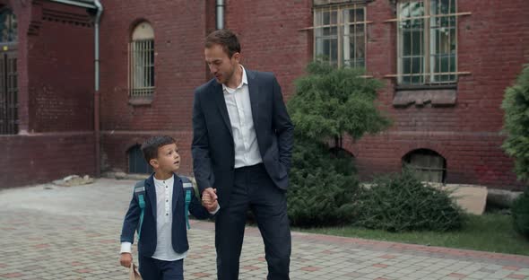 Cheerful Man in Suit and Litlle Son with Bag Holding Hand in Hand While Walking and Talking. Father