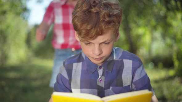 Rack Focus From Concentrated Teenage Boy Reading Book in Sunny Park to Cheerful Little Brother