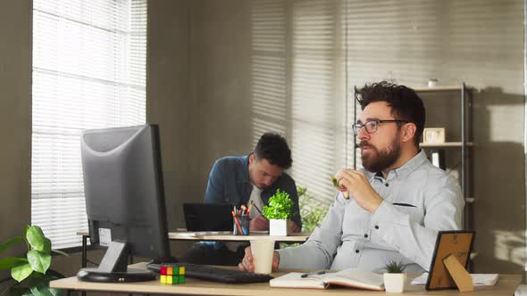 Young Man in Glasses and Casual Outfit Eating Sandwich While Working in Office