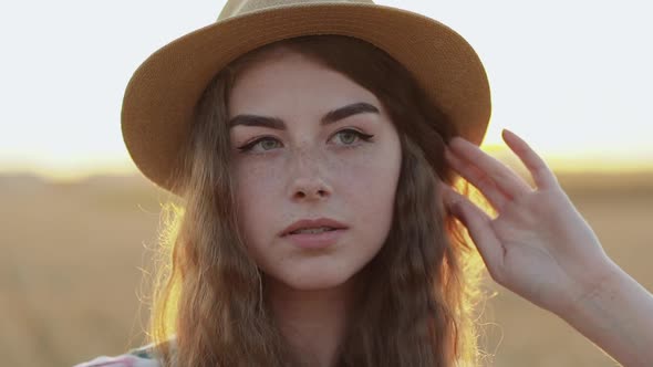 Girl with Freckles in Hat Touches Hair and Looks Aside in Field on Sunset