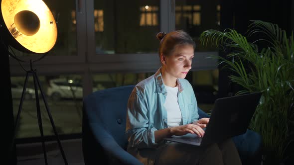 Woman is Sitting in the Armchair and Working on a Laptop at Night or Texting Someone